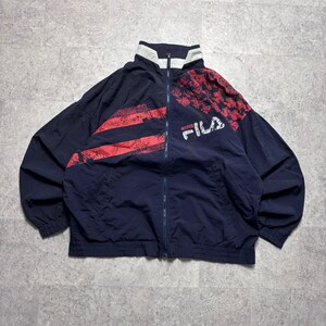 May include: Navy blue zip-up jacket with red and white accents. The jacket features a high collar and the brand name "FILA" in white. The design includes red stripes and a patterned section on the upper right shoulder.