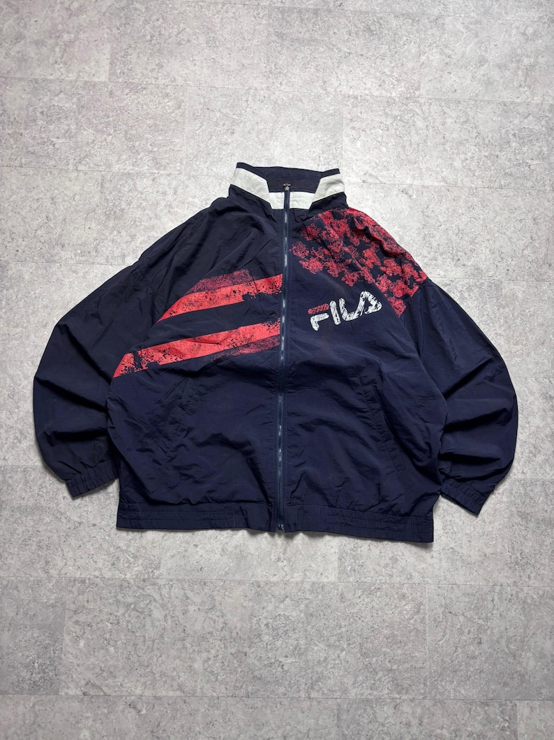 May include: Navy blue zip-up jacket with red and white accents. The jacket features a high collar and the brand name "FILA" in white. The design includes red stripes and a patterned section on the upper right shoulder.
