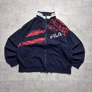 May include: Navy blue FILA track jacket with red and white accents. The jacket features a full zipper, a high collar, and the FILA logo in white. The design includes red stripes and a red and white pattern on the shoulders.