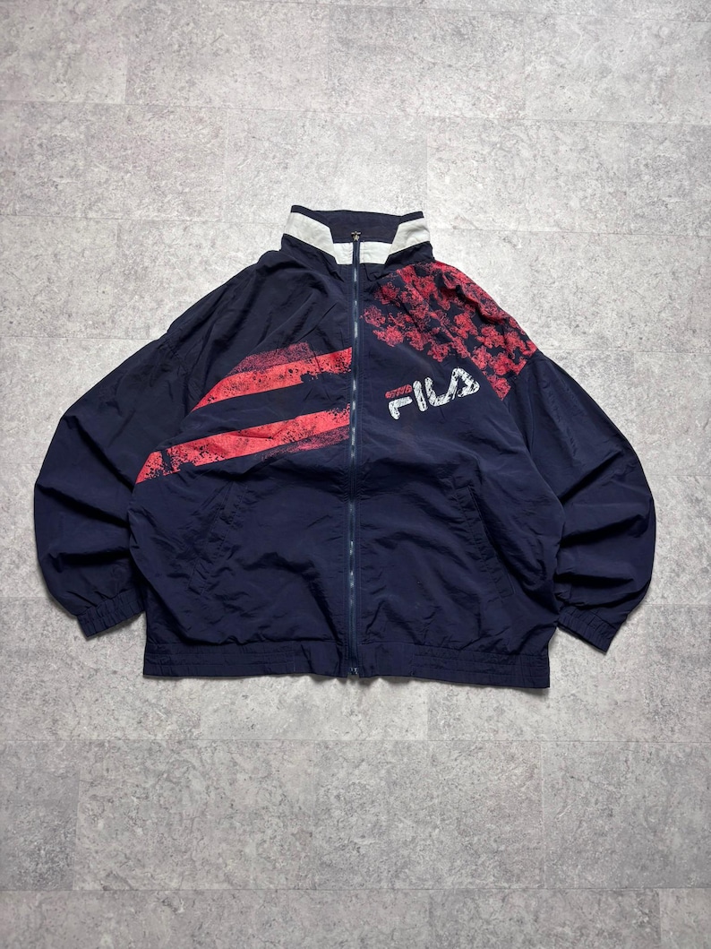 May include: Navy blue FILA track jacket with red and white accents. The jacket features a full zipper, a high collar, and the FILA logo in white. The design includes red stripes and a red and white pattern on the shoulders.