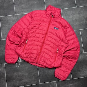 Könnte beinhalten: Eine leuchtend pinke Patagonia Steppjacke mit durchgehendem Frontreißverschluss, hohem Kragen und zwei seitlichen Reißverschlusstaschen. Die Jacke hat horizontale Steppnähte und ein kleines Patagonia Markenlogo auf der Brust.