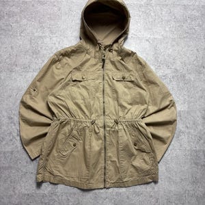 Könnte beinhalten: Eine beigefarbene Utility-Jacke mit Kapuze und Kordelzug, durchgehendem Frontreißverschluss und zwei Brusttaschen mit Druckknöpfen. Die Jacke verfügt über eine Kordelzug-Taille, zwei untere Taschen mit Druckknöpfen und lange Ärmel mit Riegeln.