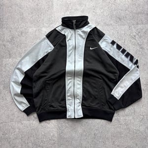 Könnte beinhalten: Schwarz-silberne Trainingsjacke mit durchgehendem Reißverschluss und hohem Kragen. Die Jacke hat silberne Einsätze an den Schultern und vorne, mit dem Nike-Logo auf der Brust. Die Ärmel haben ein Blockbuchstaben-Design.