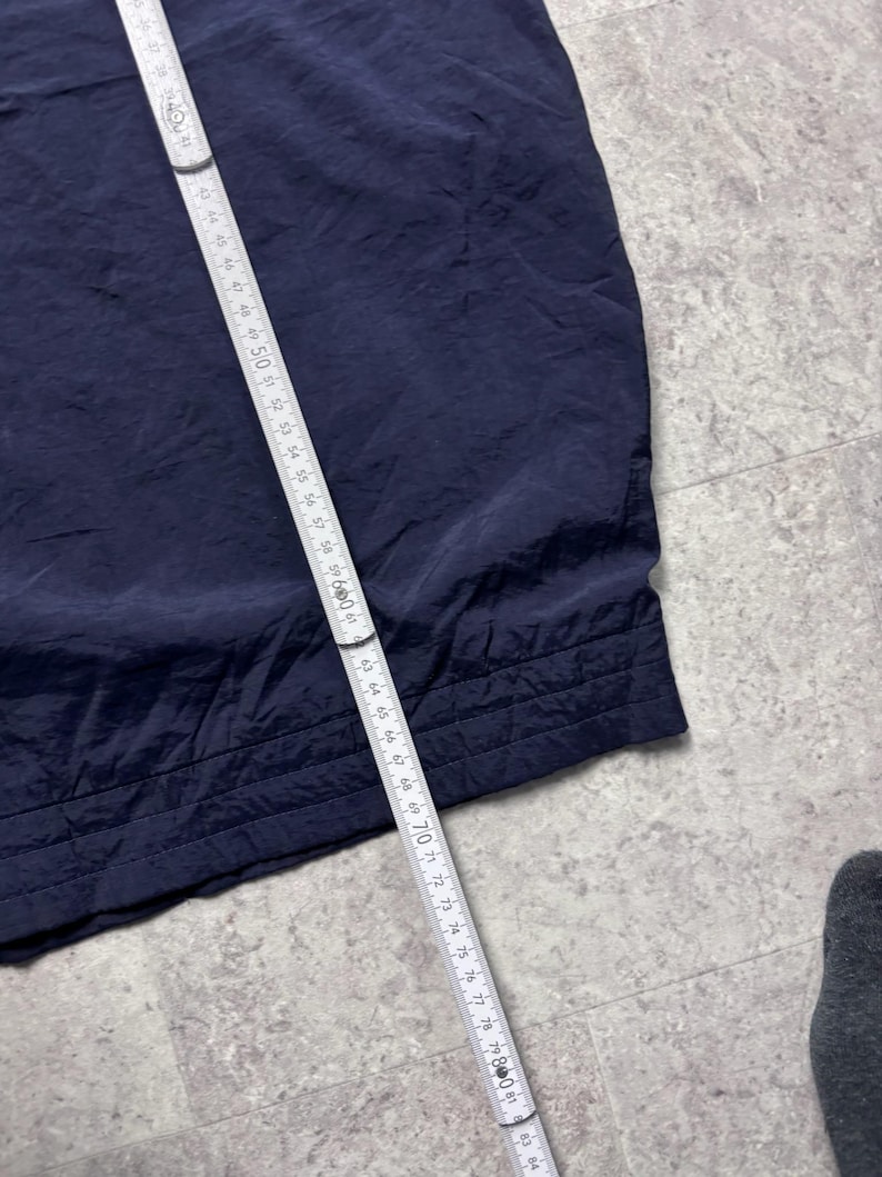 May include: A navy blue fabric, possibly clothing, is measured with a white and silver measuring tape. The tape shows measurements up to 84 inches. The fabric has a textured appearance and a stitched hem.