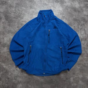 Könnte beinhalten: Eine leuchtend blaue Fleecejacke mit durchgehendem Frontreißverschluss, hohem Kragen und drei Reißverschlusstaschen. Das Kleidungsstück verfügt über ein kleines, schwarzes, aufgesticktes Adidas-Logo auf der linken Brust mit passenden Reißverschlussanhängern.