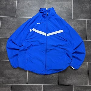 Può includere: Una giacca a vento Nike blu brillante con cerniera, caratterizzata da un colletto alto, maniche lunghe e una striscia bianca a forma di chevron sul petto. Un piccolo logo Nike swoosh bianco è ricamato sulla parte superiore destra del petto.