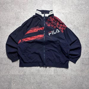 May include: Navy blue FILA track jacket with red accents. The jacket features a full zipper, a white collar, and red stripes and a red pattern on the upper right shoulder. The FILA logo is printed in white.