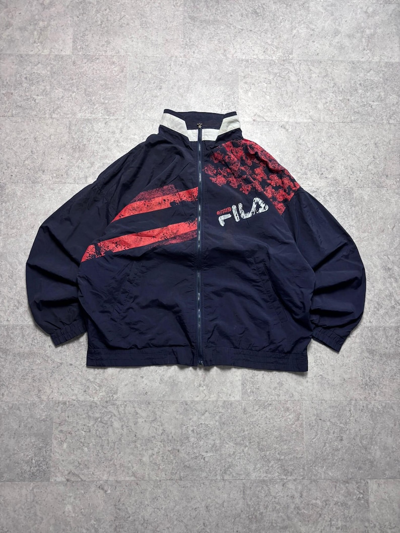 May include: Navy blue FILA track jacket with red accents. The jacket features a full zipper, a white collar, and red stripes and a red pattern on the upper right shoulder. The FILA logo is printed in white.
