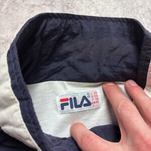 May include: Close-up of a navy blue and white Fila jacket collar. The collar is white with navy blue trim. A Fila tag is visible with the brand logo and size information.