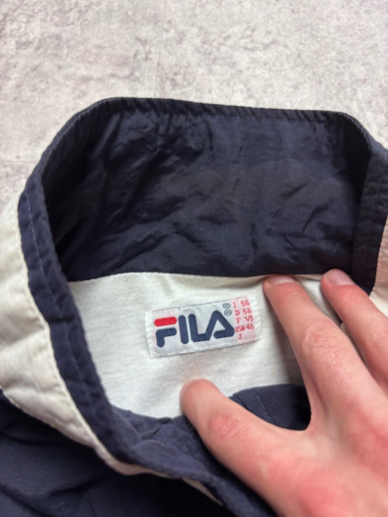 May include: Close-up of a navy blue and white Fila jacket collar. The collar is white with navy blue trim. A Fila tag is visible with the brand logo and size information.