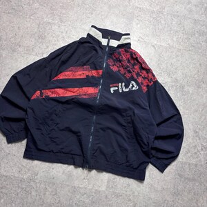 May include: Navy blue vintage Fila track jacket with red accents. The jacket features a full zipper, a white collar, and the Fila logo on the chest. The design includes red stripes and a checkered pattern on the shoulders.