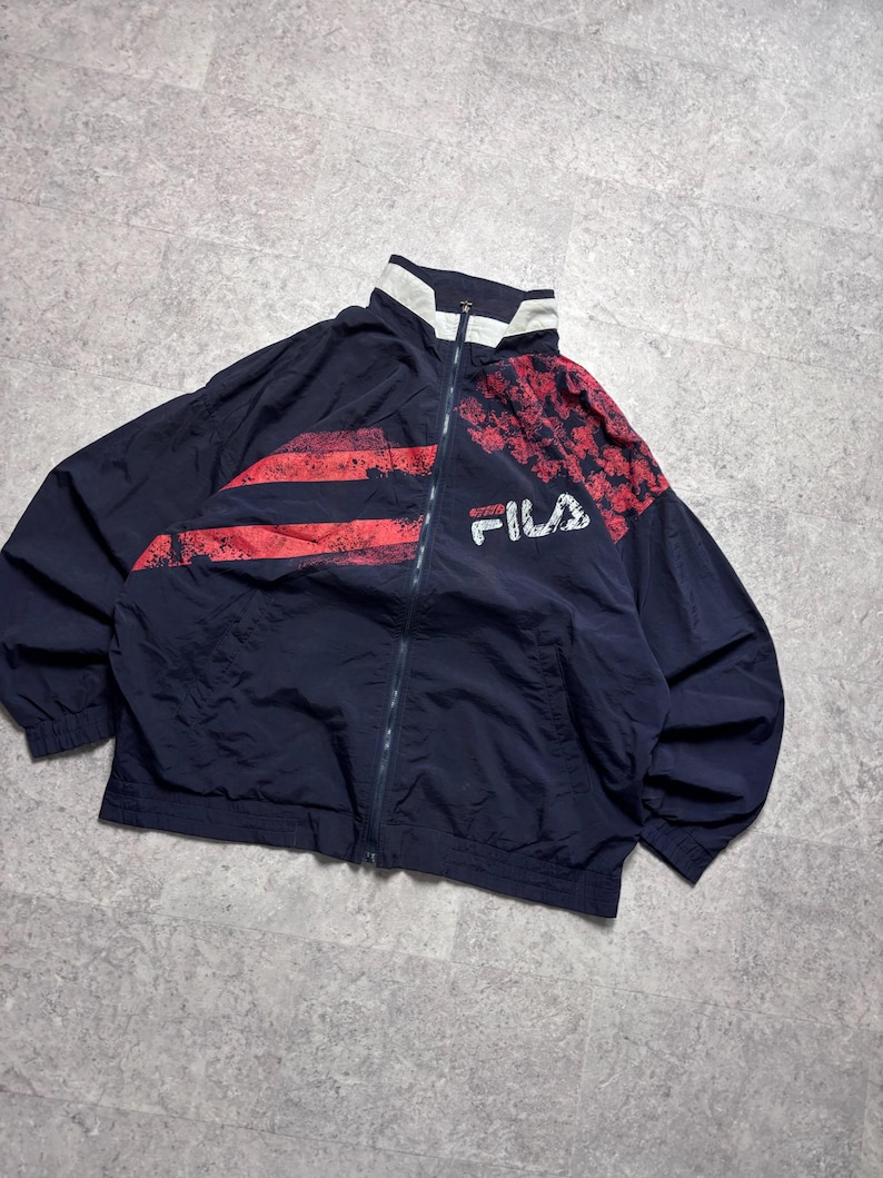 May include: Navy blue vintage Fila track jacket with red accents. The jacket features a full zipper, a white collar, and the Fila logo on the chest. The design includes red stripes and a checkered pattern on the shoulders.