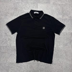Könnte beinhalten: Ein schwarzes kurzärmeliges Poloshirt mit weiß abgesetztem Kragen, einer Knopfleiste mit drei Knöpfen und einem Stone Island Markenaufnäher auf der linken Brust. Das Shirt hat ein weißes Markenetikett am Innenkragen und weiße Ärmelabschlüsse.