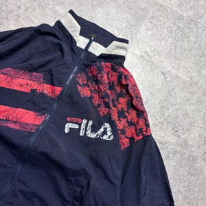 May include: Navy blue vintage Fila track jacket with red accents. The jacket features a white collar and the Fila logo in white. The design includes red splatter and patterned details on the sleeves.