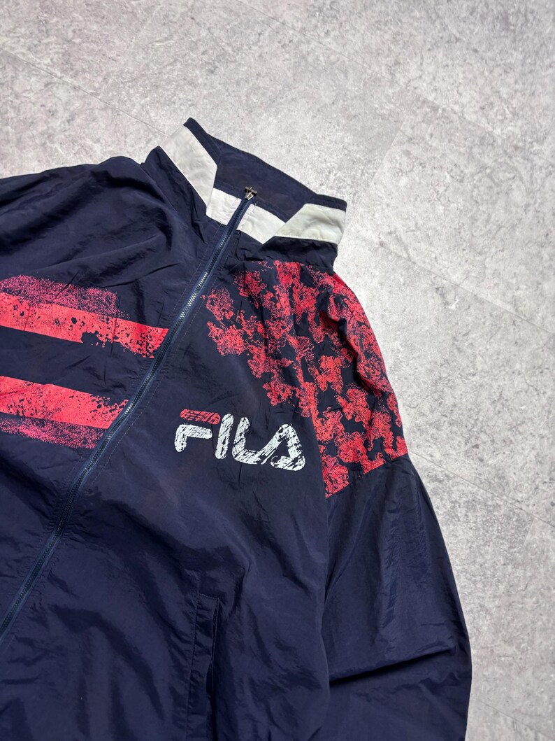 May include: Navy blue vintage Fila track jacket with red accents. The jacket features a white collar and the Fila logo in white. The design includes red splatter and patterned details on the sleeves.