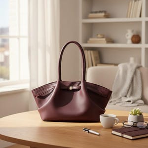 May include: A burgundy leather tote bag with a structured design and a top handle sits on a wooden table. A small gold-colored clasp secures the bag's front. A white coffee cup, a pen, a notebook, and glasses are also on the table.
