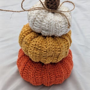 May include: A stack of three crocheted pumpkins in cream, mustard yellow, and burnt orange. Each pumpkin features a ribbed texture and a brown crocheted stem. A thin piece of natural twine is tied in a bow around the top pumpkin stem.