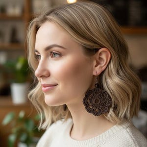 May include: A dark brown circular crochet earring featuring an intricate floral-inspired pattern with a central openwork design. The earring is suspended from a small dark bead and a metal hook, showcasing detailed needlework and texture.
