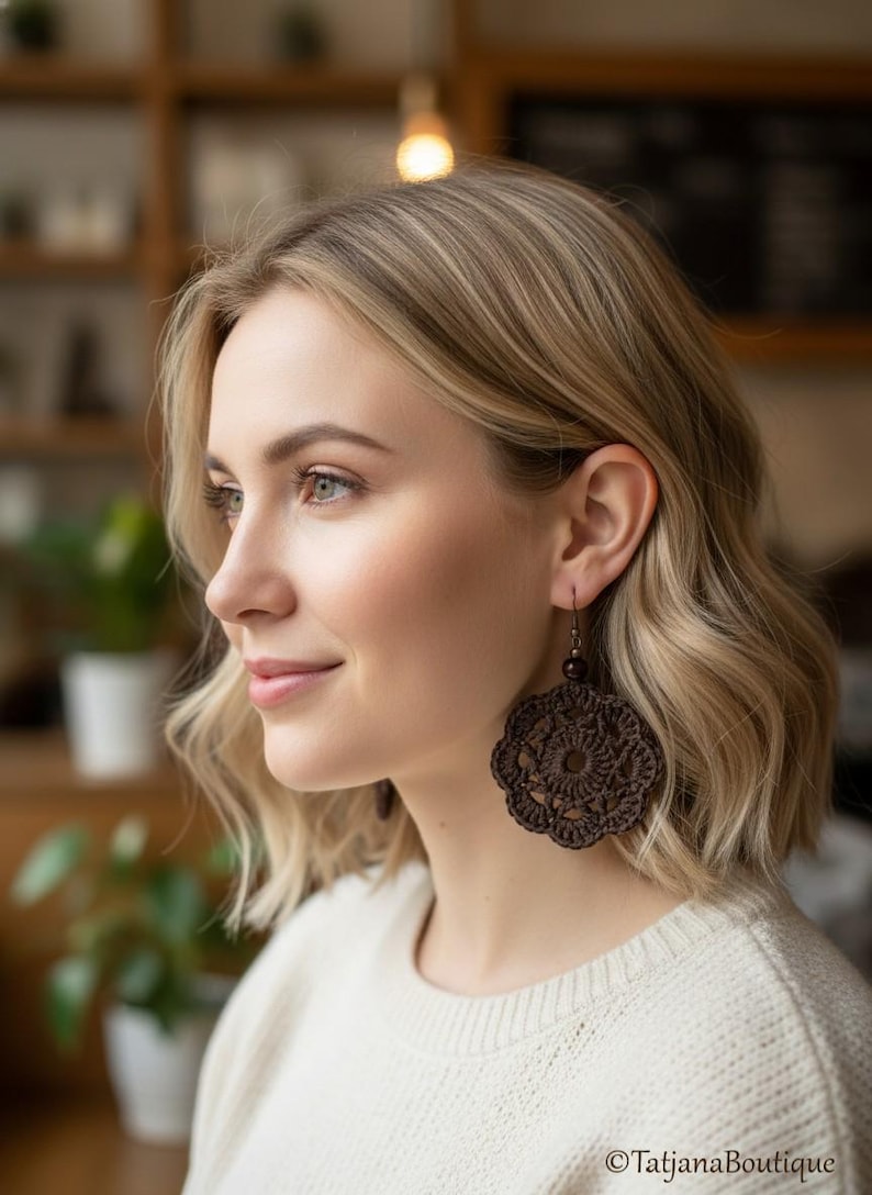 May include: A dark brown circular crochet earring featuring an intricate floral-inspired pattern with a central openwork design. The earring is suspended from a small dark bead and a metal hook, showcasing detailed needlework and texture.
