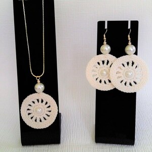 May include: A set of crocheted jewelry featuring a necklace and earrings. The necklace and earrings are white with a pearl accent in the center of each piece. The necklace has a gold chain.