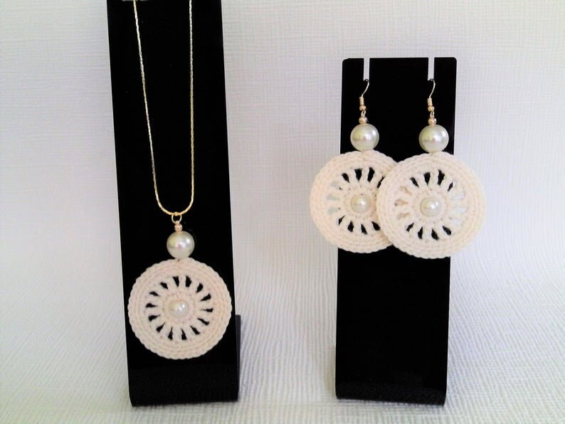May include: A set of crocheted jewelry featuring a necklace and earrings. The necklace and earrings are white with a pearl accent in the center of each piece. The necklace has a gold chain.