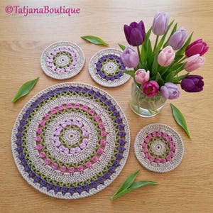 May include: Handmade crochet placemats and coasters in beige, purple, pink, and green. The placemats and coasters have a circular design with a floral pattern. A vase of tulips sits next to the placemats and coasters. The text "Tatjana Boutique" is in the top left corner.