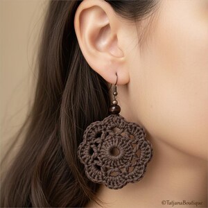 May include: A dark brown crochet earring featuring a circular, flower-like motif with an openwork pattern. The earring is suspended from a metal hook and accented with a small, dark brown spherical bead at the top of the crocheted element.