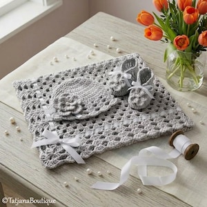 Può includere: Un set per neonati all'uncinetto grigio composto da coperta, cappello e scarpine, tutti adornati con nastri e fiocchi bianchi. Il set è esposto su una superficie di legno con perle sparse e un vaso di tulipani arancioni.