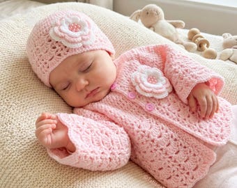 May include: A baby wearing a pink crochet hat and jacket set, both adorned with white and pink flower appliqués. The set includes a matching hat and a button-up jacket. The baby is lying on a cream-colored surface.