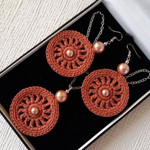 May include: A set of crocheted earrings with a circular design and a pearl accent. The earrings are displayed in a black box with a white interior.