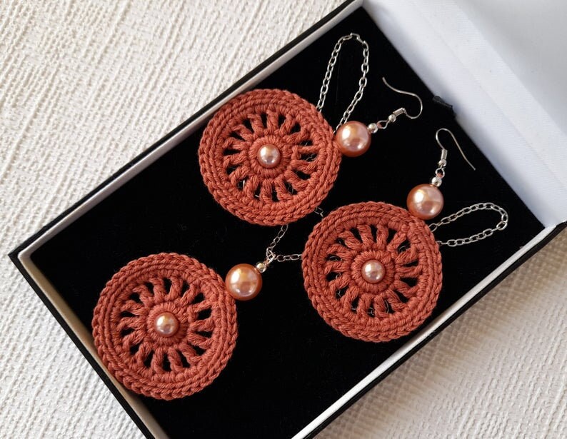 May include: A set of crocheted earrings with a circular design and a pearl accent. The earrings are displayed in a black box with a white interior.