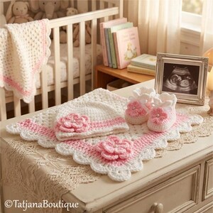 May include: A handmade baby set in white and pink, including a blanket, hat, and booties. The set features a floral design and is displayed with a framed ultrasound image, suggesting a baby shower or nursery theme.