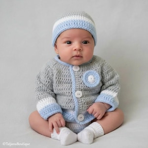 May include: A baby wearing a handmade crochet outfit. The set includes a gray cardigan with white and light blue accents, matching hat, and white socks. The cardigan has large white buttons and a decorative blue and white flower.