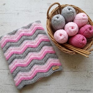 Puede incluir: Una manta de ganchillo hecha a mano con un patrón ondulado en tonos rosa y gris. Una cesta de mimbre contiene varias madejas de hilo en colores a juego. La etiqueta de la manta dice "Handmade with love".
