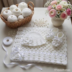 May include: A white crochet baby set, featuring a blanket, hat, and booties, decorated with satin ribbons and pearl details. A basket of white yarn balls and a bouquet of pink and white flowers are also visible. The set is designed for a baby.