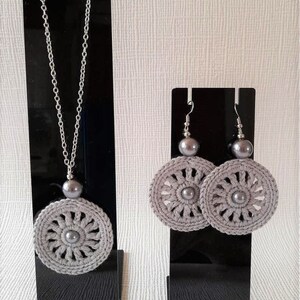 May include: A set of gray crocheted jewelry featuring a necklace and earrings. The necklace has a silver chain and a gray crocheted pendant with a pearl in the center. The earrings are silver hooks with gray crocheted circles and pearls in the center.