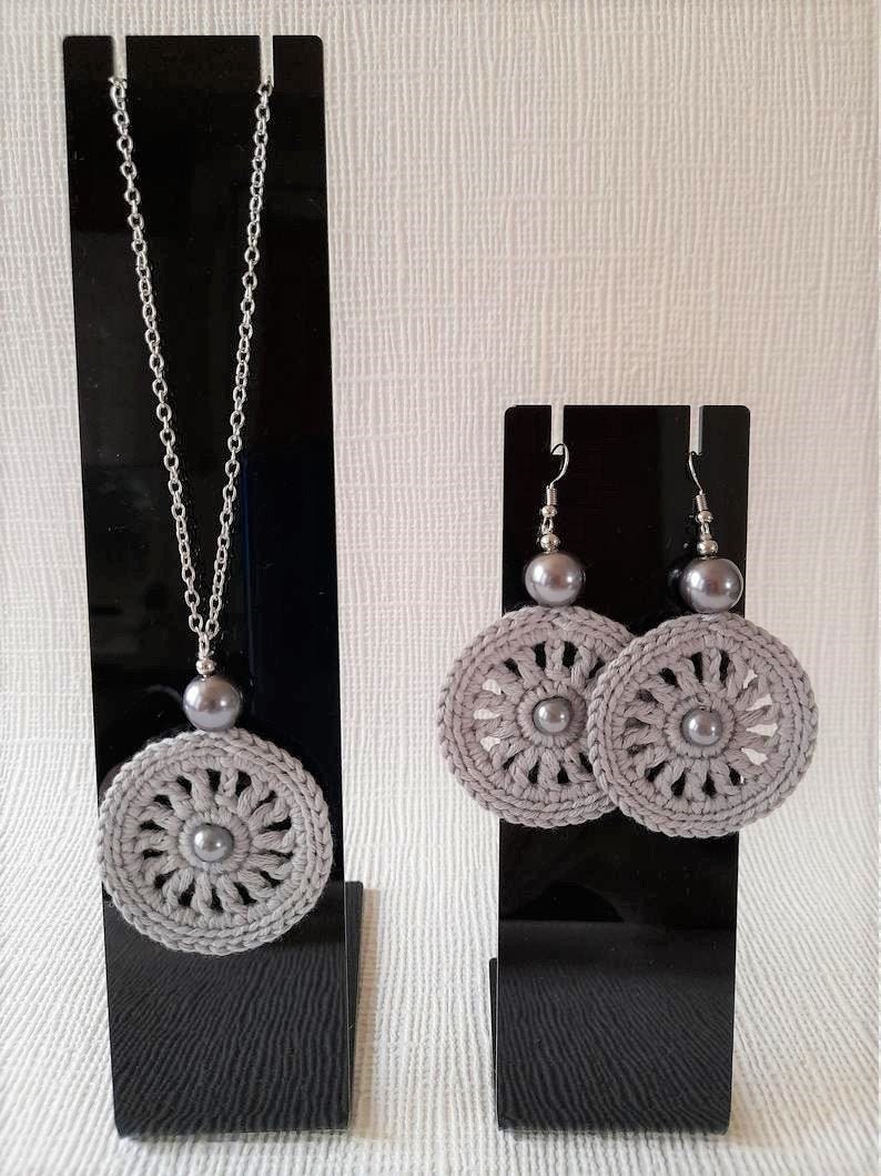 May include: A set of gray crocheted jewelry featuring a necklace and earrings. The necklace has a silver chain and a gray crocheted pendant with a pearl in the center. The earrings are silver hooks with gray crocheted circles and pearls in the center.