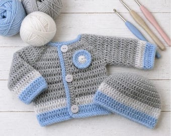 May include: A hand-crocheted gray cardigan with light blue and white striped cuffs and trim, featuring three white buttons and a circular blue and white flower accent, paired with a matching gray, white, and blue striped beanie hat.