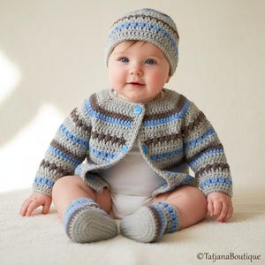 May include: A baby wearing a handmade crochet outfit. The set includes a gray, blue, and brown striped cardigan, matching hat, and booties. The cardigan has three blue buttons. The baby is sitting on a white surface.