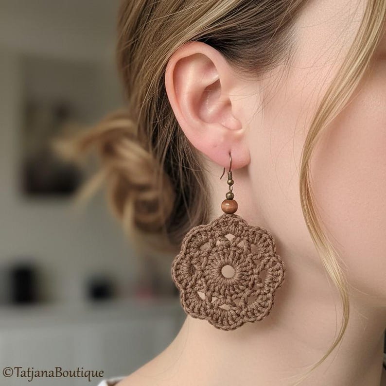 May include: A brown crochet earring featuring a circular, flower-like motif with a central open hole. The earring is suspended from a bronze-toned metal hook and includes a small, round, matching brown wooden bead above the crocheted piece.