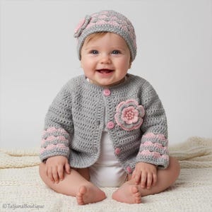 May include: A gray crochet baby cardigan and matching hat set with pink accents. The cardigan features a large pink flower and pink buttons. The hat has a flower and scalloped pink trim. A sweet baby outfit.
