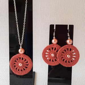 May include: A set of crocheted earrings and a necklace with a peach-colored pearl accent. The earrings and necklace feature a circular design with a crocheted pattern in a burnt orange color.