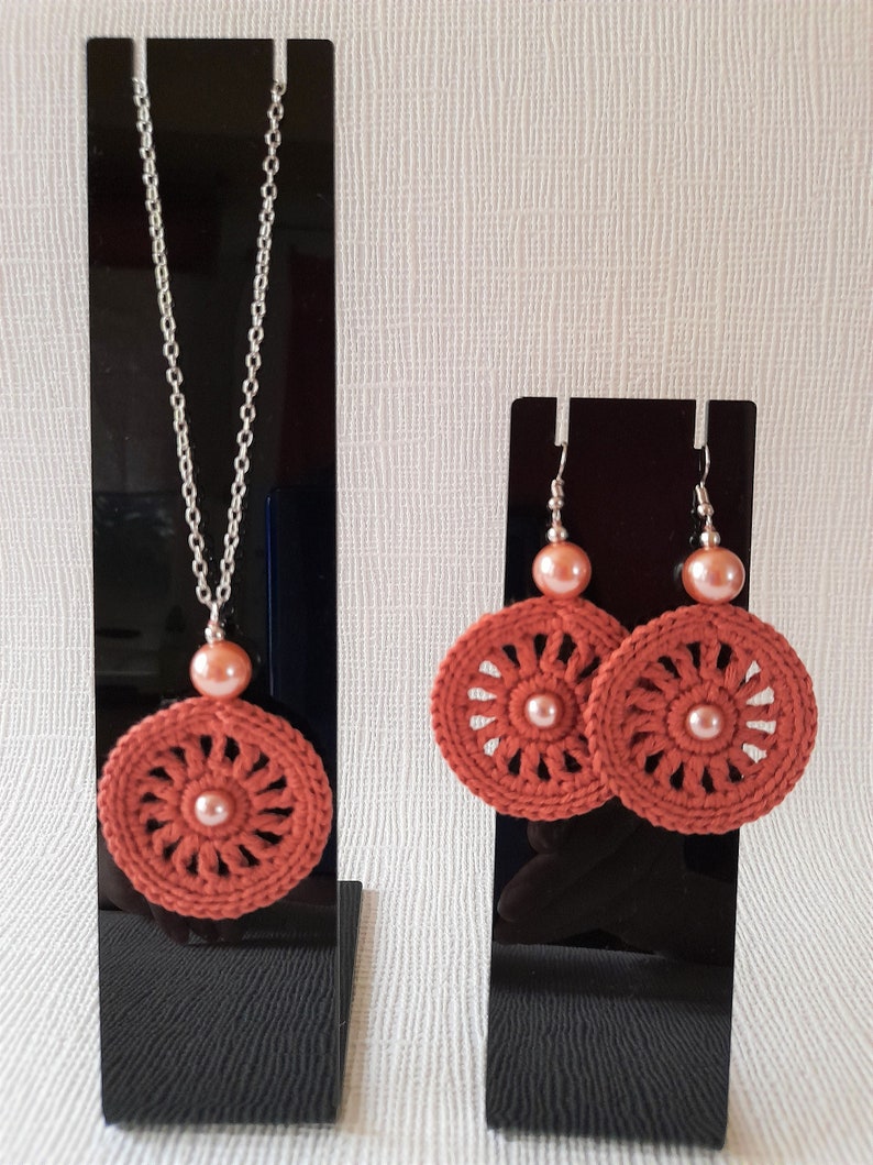 May include: A set of crocheted earrings and a necklace with a peach-colored pearl accent. The earrings and necklace feature a circular design with a crocheted pattern in a burnt orange color.