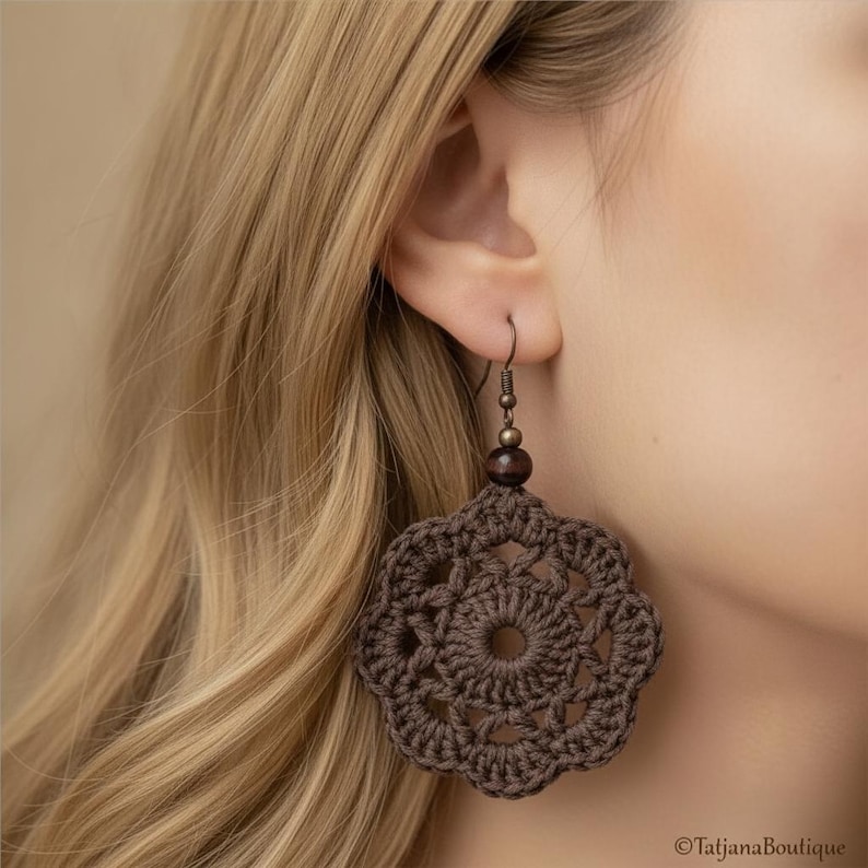 May include: A dark brown crochet earring featuring a circular, flower-like motif with an intricate openwork pattern. The crochet piece is suspended from a metallic hook by a small, dark brown spherical bead, creating a dangling design.
