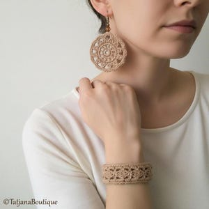 May include: Tan crochet earrings and bracelet set. The earrings are large, circular, and feature a detailed openwork design. The matching bracelet is wide and also has an openwork pattern. The jewelry is handmade and appears to be made of a natural fiber.