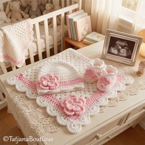 May include: A handmade baby set in white and pink, including a blanket, hat, and booties, all with a floral design. The set is displayed on a white surface with a framed ultrasound image and a crib in the background.