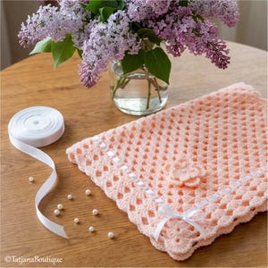 May include: A peach-colored, crocheted blanket with a scalloped edge and white ribbon accents. A small crocheted flower is attached to the blanket. A roll of white ribbon and a bouquet of purple lilacs in a glass vase are also present.
