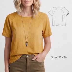 Può includere: Top color giallo senape a maniche corte con maniche a raglan. È visibile una lunga collana dorata con un ciondolo a forma di piuma. Il testo "Taglie: 32 - 56" si trova nell'angolo in basso a destra. Un disegno al tratto del top è a destra.