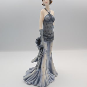 May include: A figurine of a person with short brown hair wearing a floor-length, light blue and grey gown with a halter neckline and a dark swirl pattern on the bodice. The figure wears long matching gloves and holds a draped fabric piece.