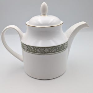 Puede incluir: Una tetera de porcelana blanca con una tapa que tiene un remate puntiagudo y un borde dorado. El cuerpo está decorado con una banda horizontal de motivos florales en color verde salvia y blanco, junto a un asa curva y un pico vertedor.